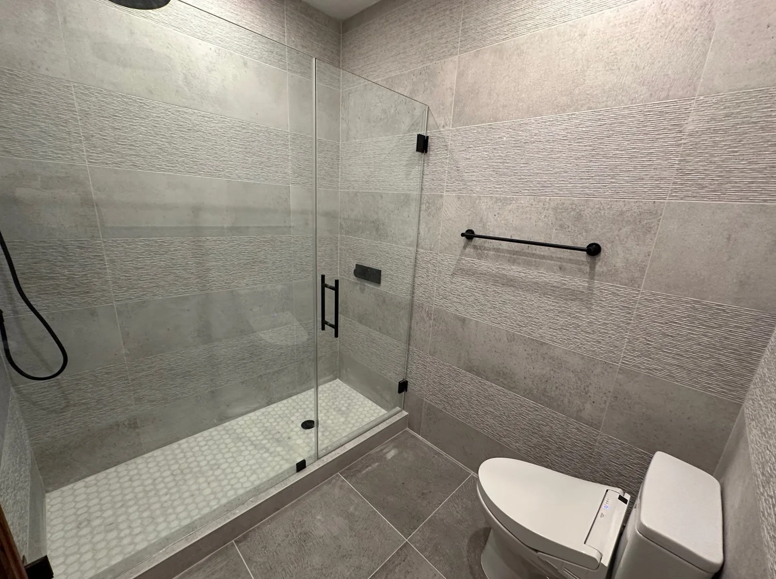 Frameless glass shower with black handle, hinges and handheld hose, black towel bar and large gray tile