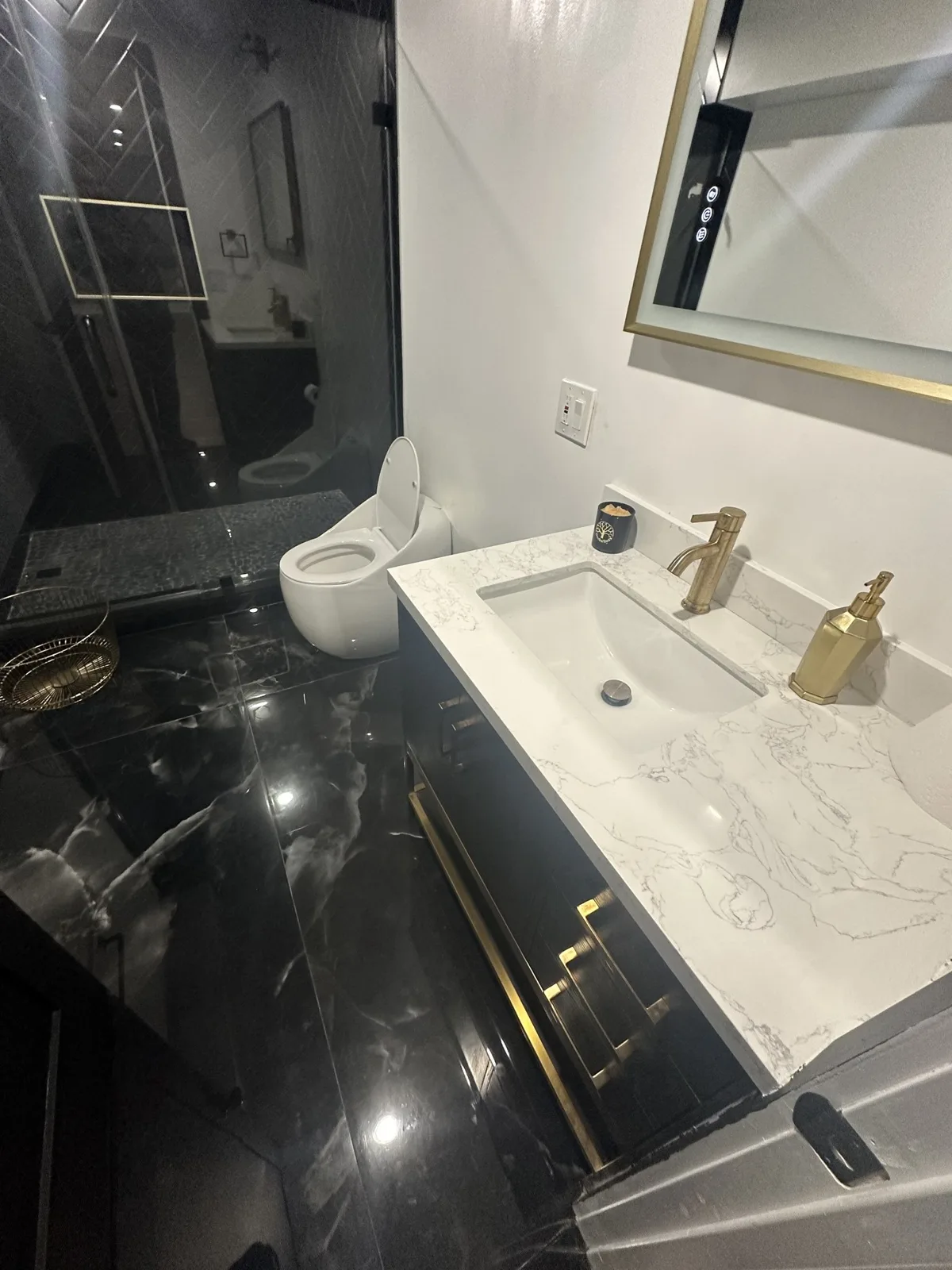 Bathroom vanity with gold faucet and gold pulls, white stone-look top, gold-framed lighted mirror and black marble-look floor tile
