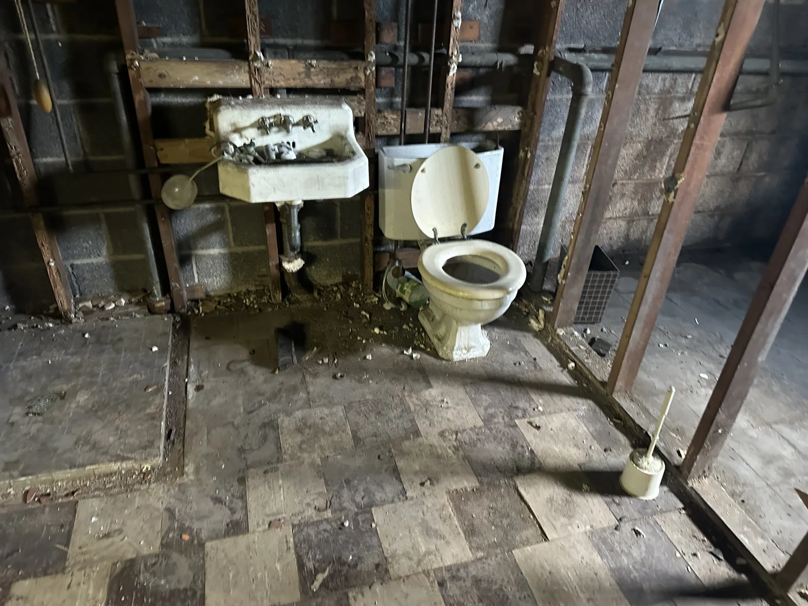 Bathroom stripped to the studs and block walls, with an old wall-hung sink, toilet and exposed pipes