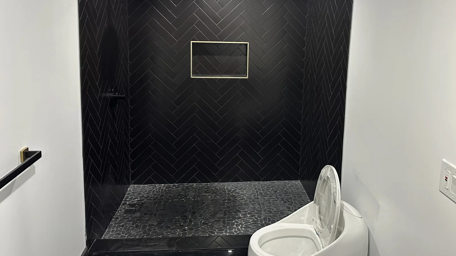 Finished walk-in shower with black herringbone wall tile, a black pebble floor and a gold-trimmed niche