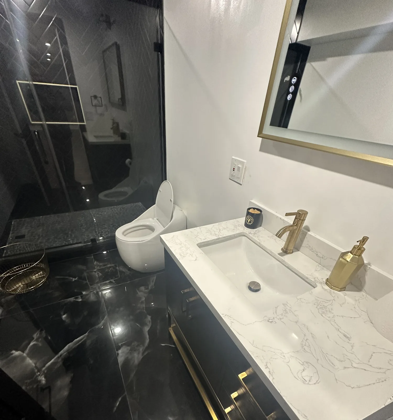 Dark vanity with gold faucet and hardware, white stone-look top, gold-framed mirror and black marble-look floor tile
