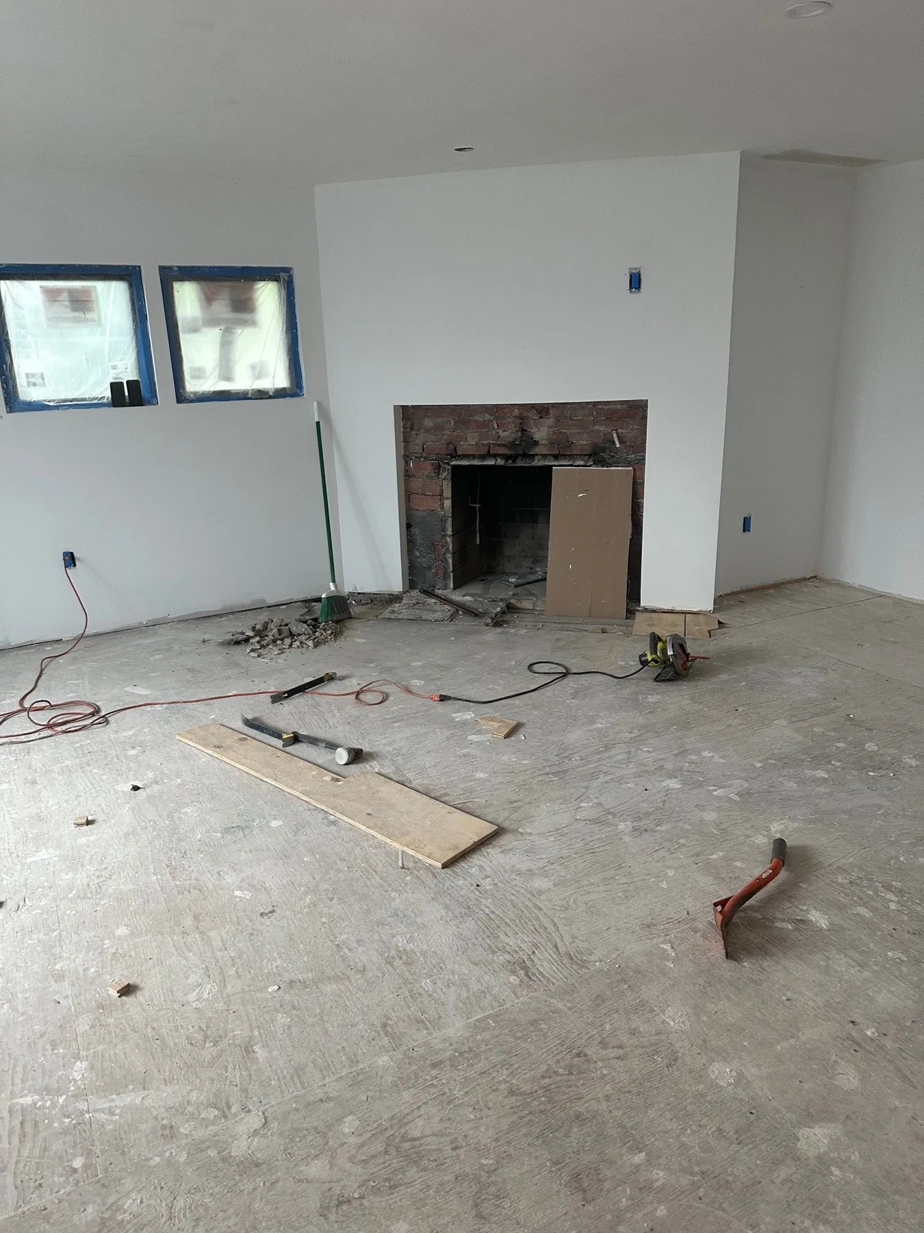 Old chipped brick fireplace opening on a freshly drywalled chimney wall, with loose masonry on the bare subfloor in front of it