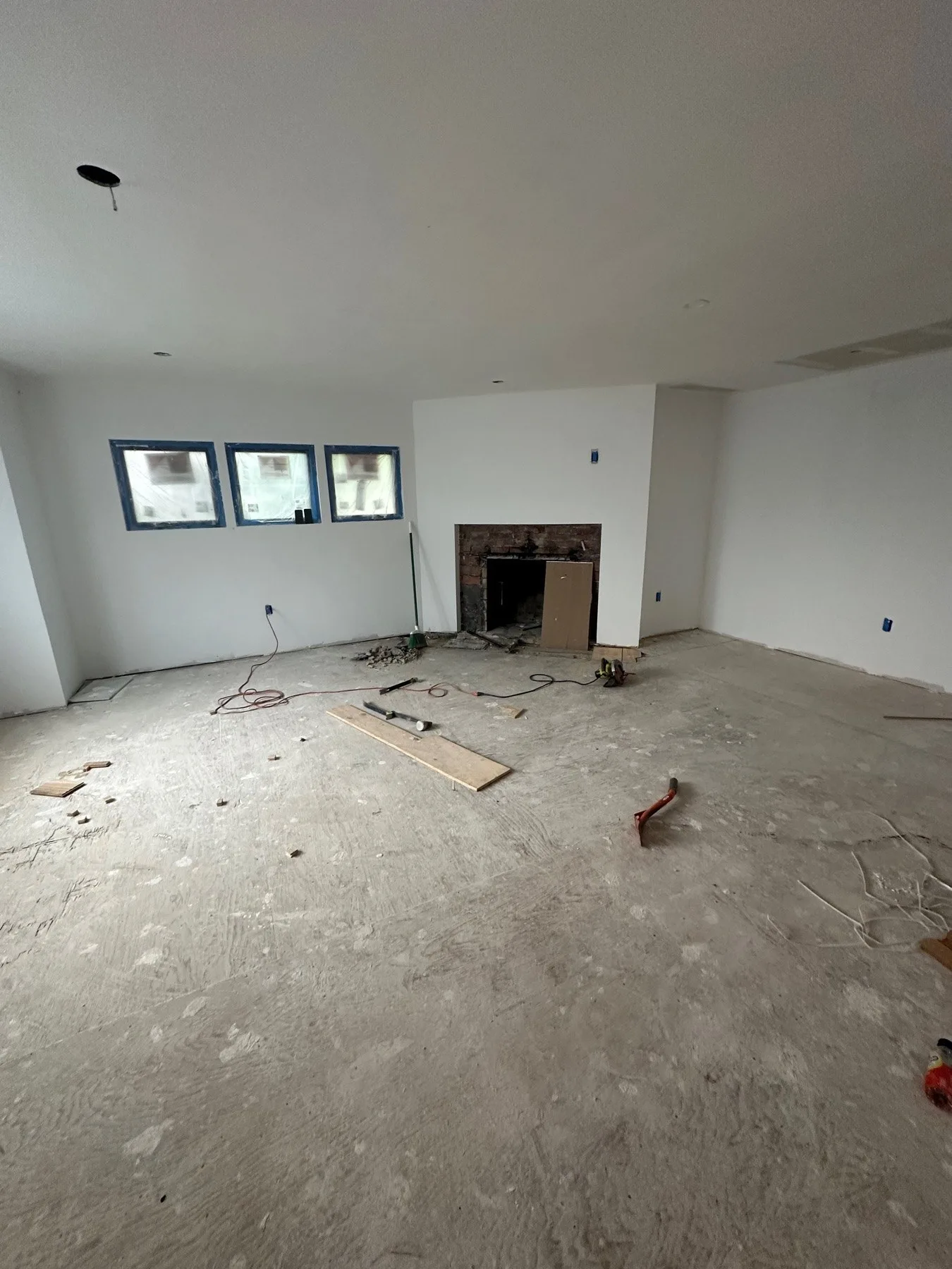 Large living room before renovation, stripped to a bare plywood subfloor with scattered debris and tools, an old brick fireplace on the far wall and a row of black-framed windows