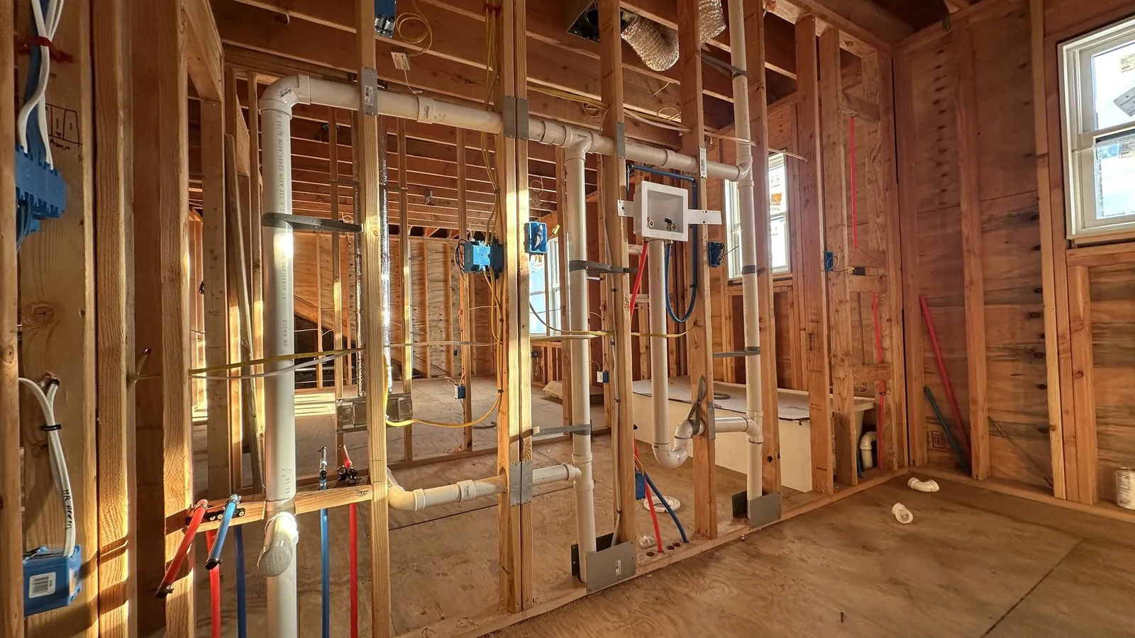 Framed interior walls during rough-in with white PVC drain and vent pipes, red and blue PEX water lines, blue electrical boxes and wiring run through wood studs