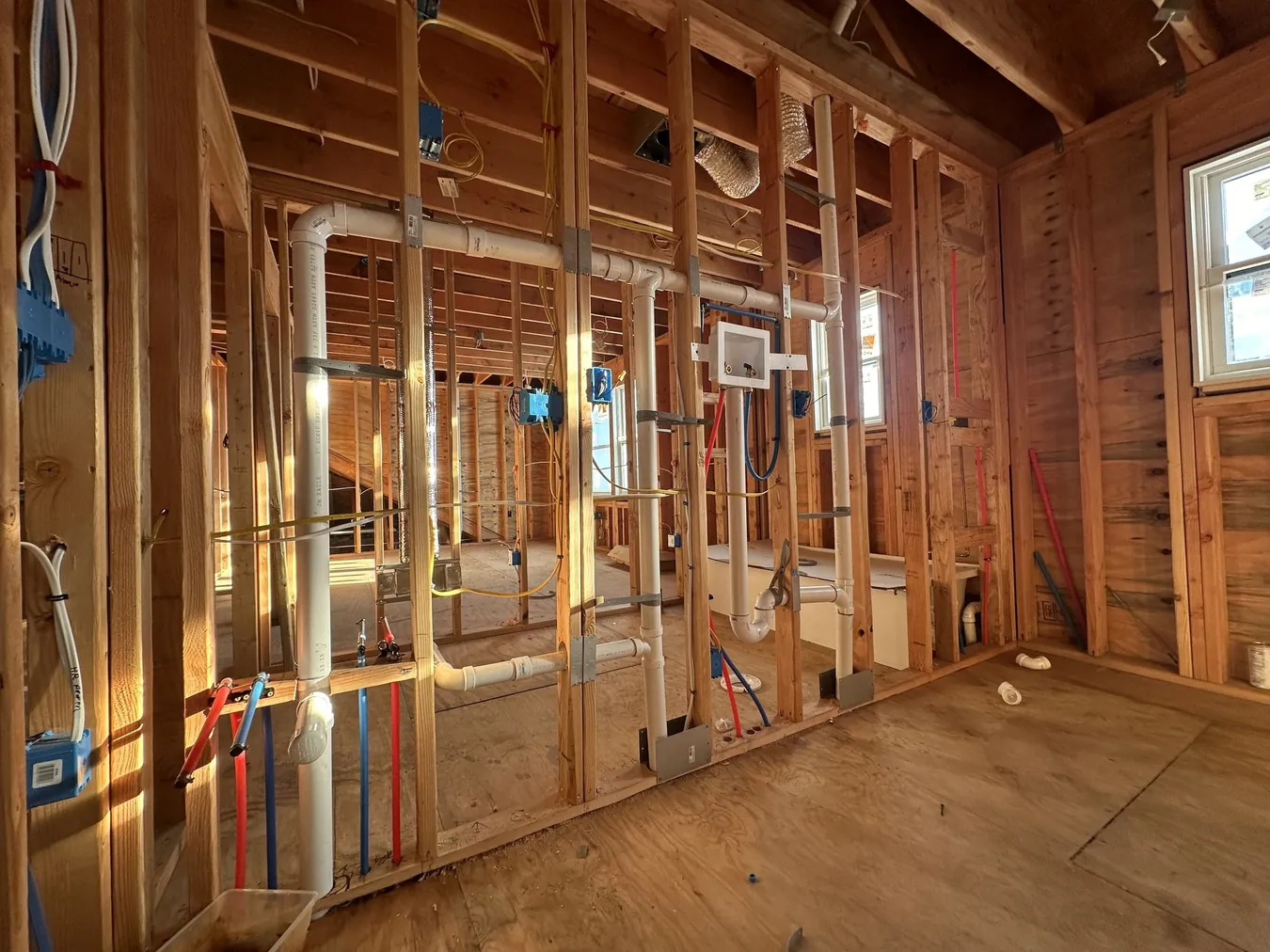 Framed interior walls during rough-in with white PVC drain and vent pipes, red and blue PEX water lines, blue electrical boxes and wiring run through wood studs