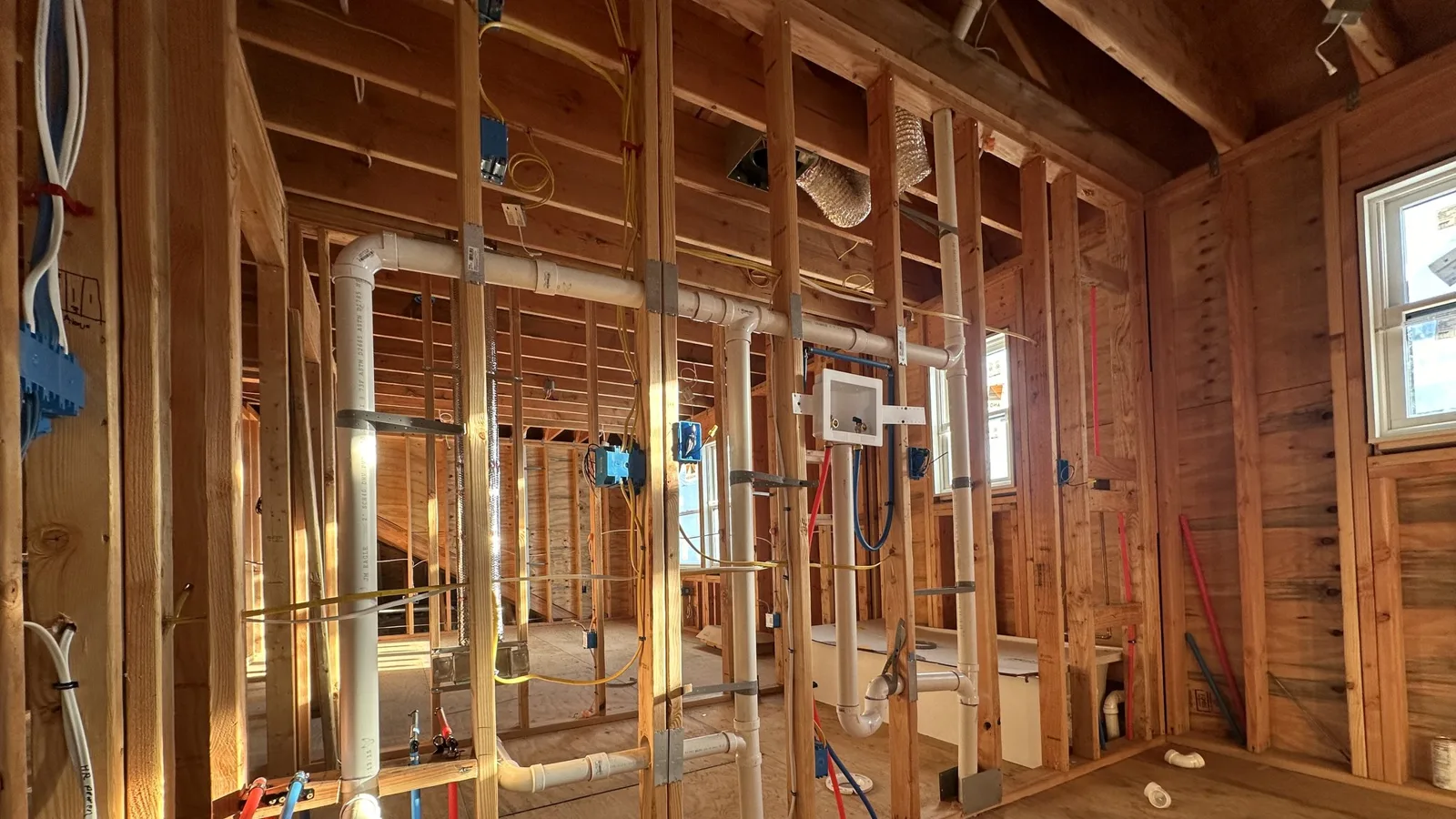 Open stud walls with PVC drain lines, red and blue PEX water lines, and blue electrical boxes before insulation