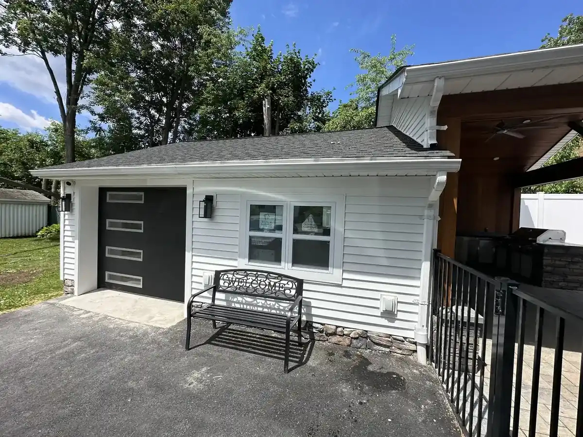Renovated detached garage with white siding, a stone skirt and a modern black glass-panel garage door, beside an ipe-clad covered patio